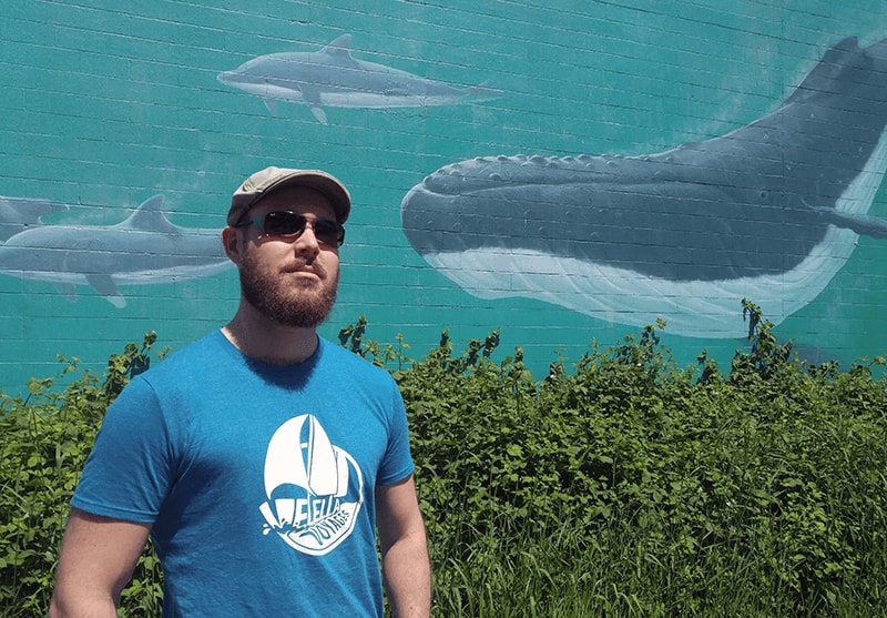 justin holmes wearing velella voyages tshirt beside mural of undersea scene with whales and dolphins by todd polich